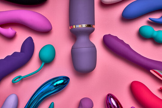 Colorful assortment of vibrators and sex toys displayed on a pink background, showcasing different shapes, sizes, and designs for adult pleasure.