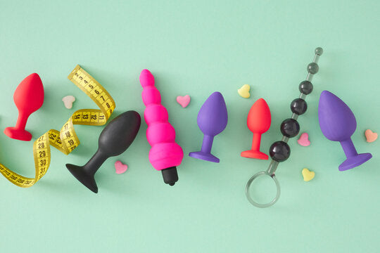 Assortiment de plugs anaux en silicone multicolore, chapelet anal et vibromasseur rose disposés sur fond vert menthe avec ruban à mesurer et petits cœurs décoratifs.