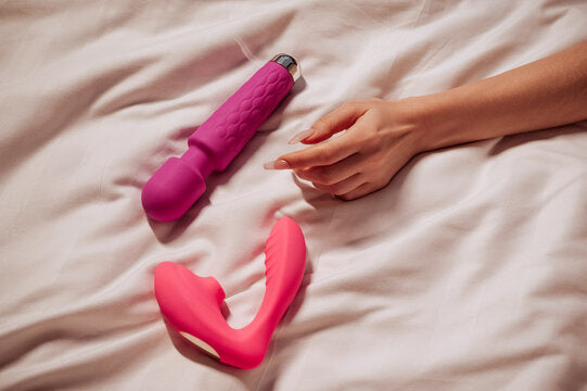 Deux sextoys roses posés sur un lit avec une main féminine, illustrant des jouets sexuels pour femme modernes et design.