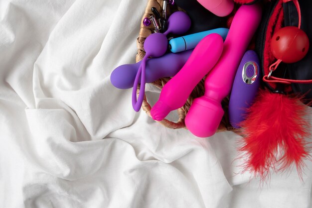 Collection de jouets sexuels colorés pour adulte sur drap blanc – vibrateurs, plugs, accessoires BDSM.