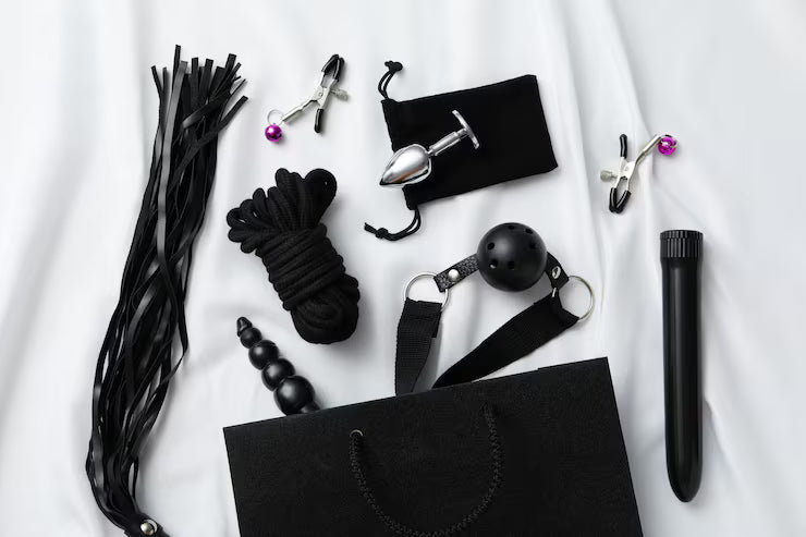 Accessoires BDSM pour adultes comprenant fouet, corde, bâillon, plug anal métallique, pinces à tétons et vibromasseur disposés sur un drap blanc.