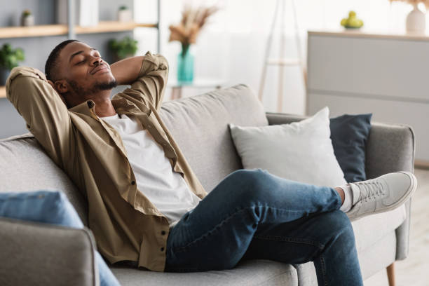 Homme détendu assis sur un canapé dans un salon moderne, les yeux fermés et les mains derrière la tête, profitant d’un moment de relaxation à la maison.