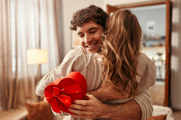 Couple heureux s’embrassant à la maison, l’homme tenant une boîte cadeau en forme de cœur rouge, célébrant un moment romantique ou la Saint-Valentin.