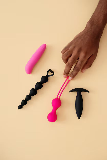 Assortiment de jouets intimes en silicone rose et noir — mini vibromasseur, chapelet anal, boules de Kegel et plug — posés sur fond beige.
