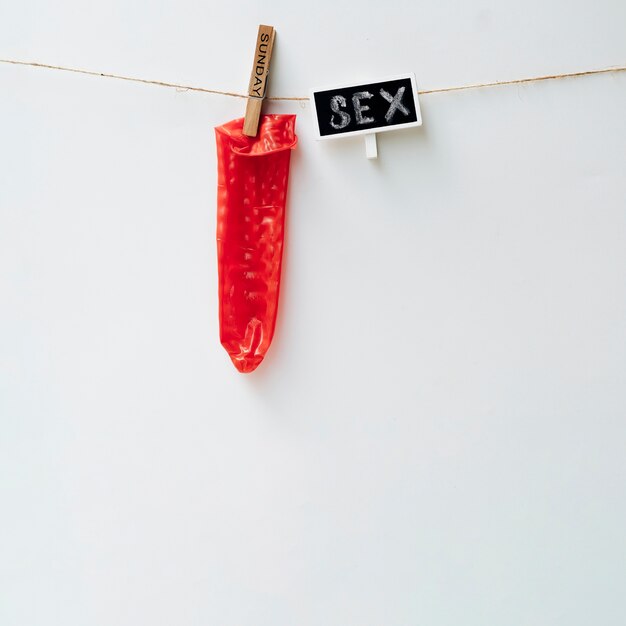 Préservatif rouge suspendu avec une pince à linge marquée 'Sunday' et une étiquette 'SEX' sur un fil, sur fond blanc.