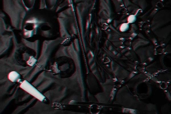 Accessoires BDSM pour adultes comprenant masque, fouet, menottes, plug anal et vibromasseur disposés sur un tissu noir.