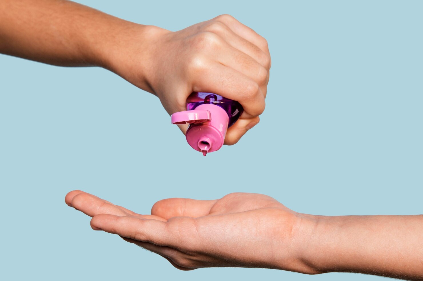 Main pressant une bouteille de gel désinfectant rose sur une paume ouverte, fond bleu clair.