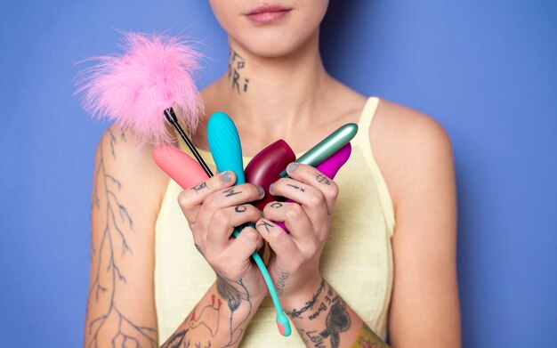 Femme tenant plusieurs jouets sexuels colorés et accessoires érotiques sur fond violet.